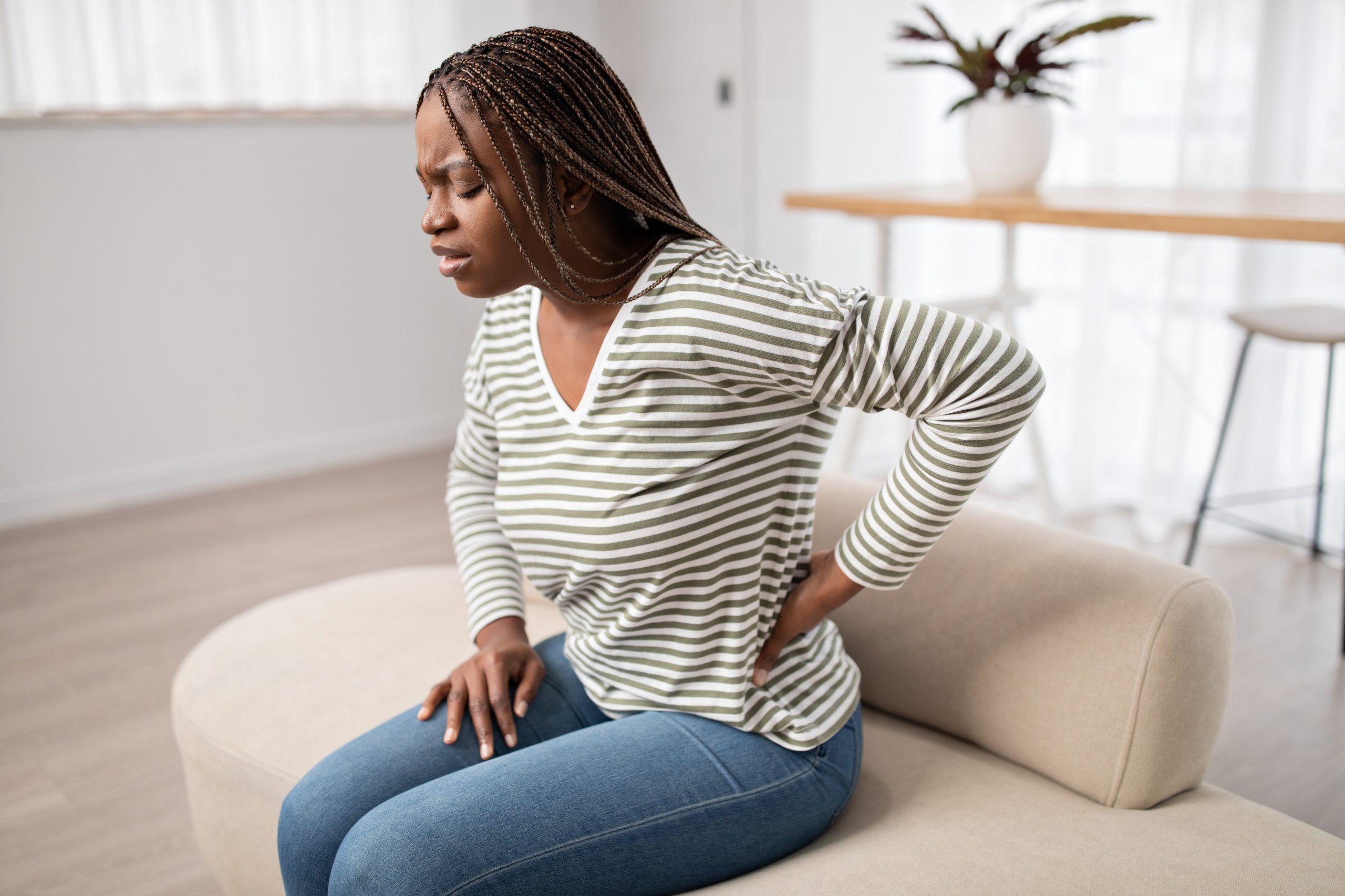 Upset young African-American long-haired woman in pain sitting on couch, touching her lower back, african american lady suffering from backache, muscle strain, period cramps.