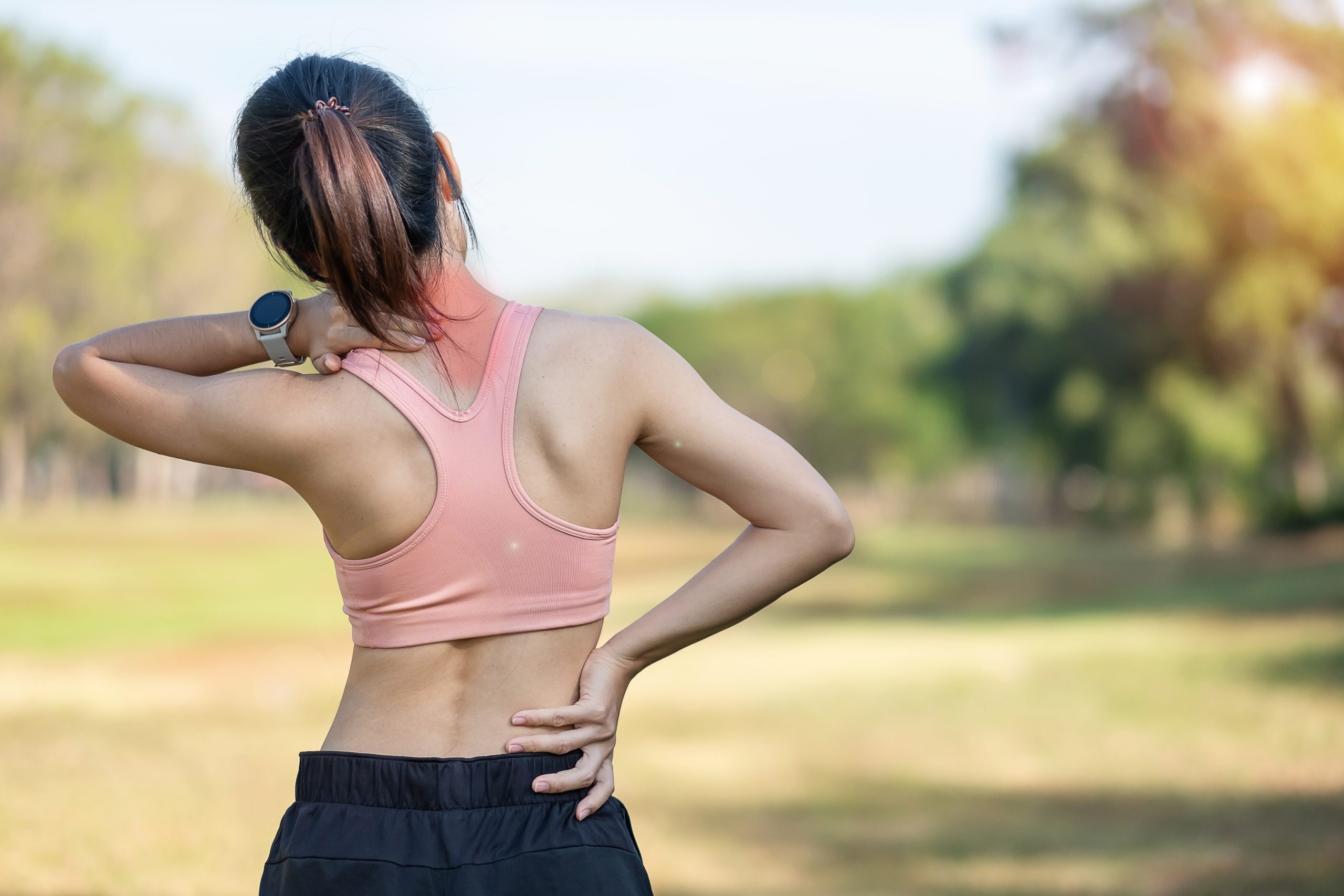 A young woman in workout clothes, holding her neck and back in pain.