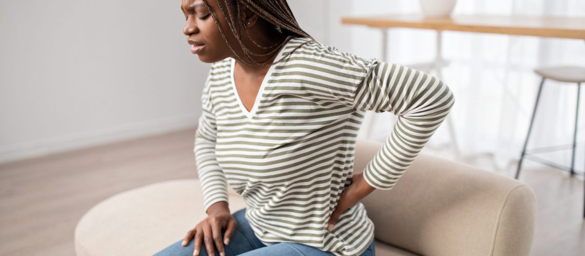 Upset young African-American long-haired woman in pain sitting on couch, touching her lower back, african american lady suffering from backache, muscle strain, period cramps.